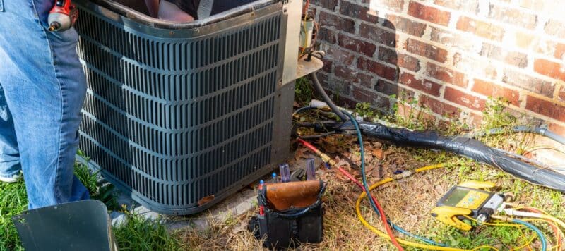 ac technician working inside an outdoor condenser unit installed outside a red brick home. tools lay on the grass nearby